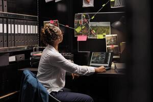 African american policewoman analyzing evidence board while solving crime. Law enforcement professional looking at clues connection scheme hanging on wall and searching insight photo