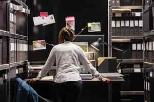 Policewoman standing near workplace and studying detective board hanging on wall. African american woman police investigator analyzing evidence and crime scene photographs connection map photo