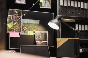 Crime evidence board with investigation process map lit with desk lamp in private investigator room. Police detective gathered information hanging on wall and table with laptop in office photo