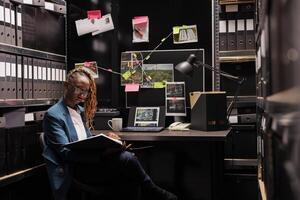 africano americano mujer sentado a detective lugar de trabajo escritorio y leyendo crimen caso archivo. privado investigador estudiando forense inspector conclusión y buscando visión a noche hora foto