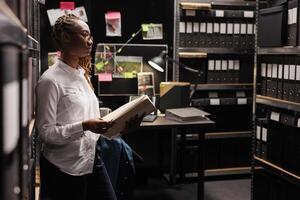 Law enforcement professional reading investigation materials in detective office room. African american woman investigator holding crime case file and looking at folders in shelf photo