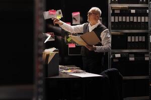 Senior police officer holding confidential files, looking at investigation board in arhive room. Elderly private detective working overhours at criminal case, checking mysterious suspect report photo