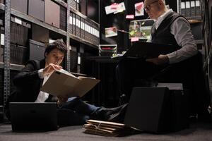 Tired police officers holding files with crime scene evidence, analyzing criminology report in arhive room. Overworked private detectives working late at night at missing person case photo