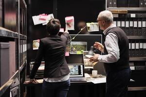 Private detectives looking at investigation board, analyzing crime scene evidence in arhive room. Elderly police officer explaining missing person report to investigator, working at criminal case photo