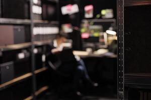 Selective focus of shelves full with evidence files, in background private investigator reading crime scene report. Police officer working late at night at missing person case in arhive room. photo