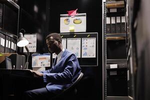 African american manager looking at bookkeeping files, analyzing accountancy documents in storage room. Corporate businessman in formal suit organizing administrative paperwork photo