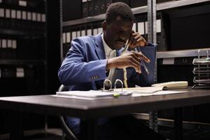 African american private detective talking with remote coworker using landline phone, discussing criminal investigation. Police officer analyzing evidence trying to solve case in arhive room photo