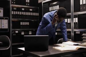 African american inspector reading police expertise, working late at night at criminal case in arhive room. Private detective analyzing crime scene evidence trying to catch mysterious suspect photo