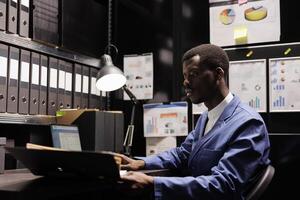 Police officer analyzing criminology report, checking crime scene evidence in arhive room. African american private detective discovery confidential information about mysterious suspect photo