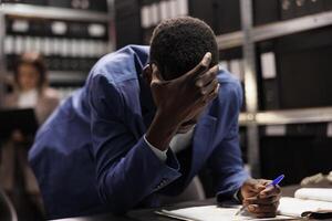 Pensive businessman trying to solve corporate report, analyzing administrative documents in storage room. African american bookkeeper searching for management files, checking bureaucracy record photo