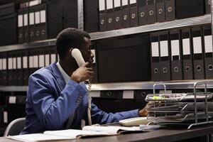 Police officer having remote conversation with inspector, discussing crime scene report using landline phone. African american detective working overhours at mysterious case in arhive room photo