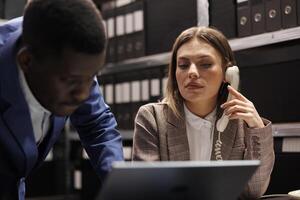 Secretary talking with bookkeeper at landline phone, working together at accountancy report in corporate depository. Diverse businesspeople analyzing administrative documents in file room photo