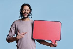 Cheerful arab man pointing at red blank banner while advertising product. Smiling person with positive facial expression holding dialog cloud for promotional message and looking at camera photo