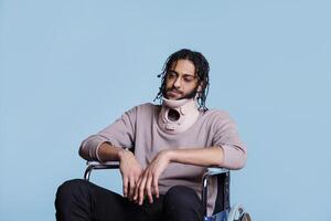 Exhausted arab man with neck brace sitting in wheelchair, healing from accident. Tired young patient wearing cervical collar protector for spinal injury recovering from trauma photo