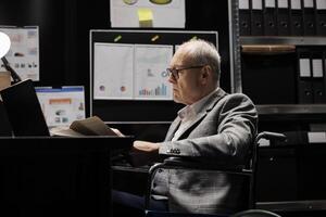 Business manager executive in wheelchair at office desk inspecting quarterly company results analysis data paperwork before consulting meeting. File cabinet office filled with folders photo