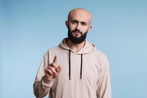 Serious arab man pointing at camera and showing one studio portrait. Young adult bald bearded arabian person raising index finger and looking with confident facial expression photo