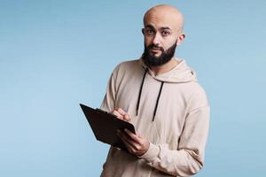 Arab man taking notes on clipboard and looking at camera. Young bald arabian person wearing casual hoodie holding checklist, planning and writing with pen studio portrait photo