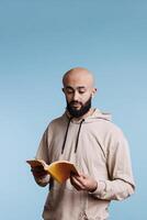 Arab man reading book paperback edition with yellow cover. Young concentrated bald bearded person in hoodie holding open notepad while standing with surprised facial expression photo