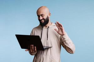 árabe hombre participación ordenador portátil mientras hablando en vídeo llamar, sonriente y ondulación Hola. alegre persona teniendo remoto comunicación utilizando portátil computadora aplicación y saludo cliente foto