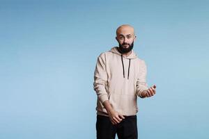 Young arab man in good mood dancing and looking at camera. Relaxed bald bearded person in casual hoodie making hands and body moves, feeling rhythm and partying studio portrait photo