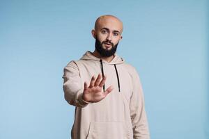 Arab man making stop gesture with hand while looking at camera with serious facial expression. Young person standing in confident pose while showing prohibition and disapproval portrait photo