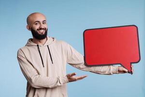 Smiling arab man blank red message frame with place for text with cheerful facial expression portrait. Carefree young person holding empty comic balloon and looking at camera photo