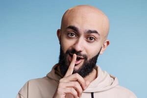 Young arab man making shh gesture with forefinger on lips and keeping silent portrait. Handsome bald bearded person showing quiet sign with finger on mouth and looking at camera photo