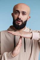 Arabian person taking time out with stop signal while showing confident facial expression portrait. Young bald bearded man making interruption gesture with hands while looking at camera photo