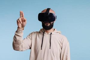 Young man in vr helmet making tap gesture with two fingers while exploring metaverse. Person in headset enjoying simulation activity and playing virtual reality games in cyberspace photo