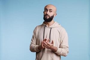 Young arab man applauding and supporting performance while looking away. Bald bearded person in casual hoodie clothes clapping with hands and showing congratulation gesture photo