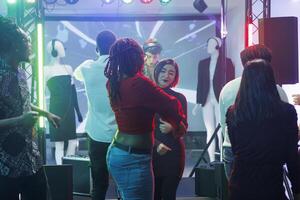 Women chatting on dancefloor while partying at electronic music discotheque in club. Diverse group of young clubbers dancing energetically and celebrating in crowded nightclub photo