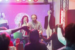 Band moving to rhythm while singing and mixing music on club stage illuminated with spotlights. Dj playing and singer with microphone dancing at discotheque in dark nightclub photo