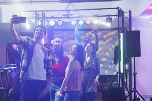 joven hombre participación teléfono inteligente y tomando grupo selfie con amigos en Club nocturno pista de baile. diverso clubbers posando para móvil teléfono foto juntos mientras bailando a club fiesta