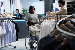 Man in wheelchair exploring clothing store and receiving guidance from mall assistant in selecting casual outfit. Shopping center customer with disability browsing through apparel rack photo