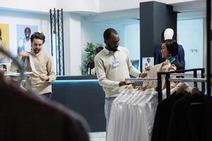 Clothing store african american worker helping asian woman to choose garment while exploring apparel rack. Boutique employee providing client support and assistance in selecting trendy outfit photo