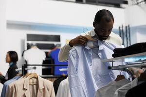 joven africano americano comprador molesto en camisa mientras elegir vestir Talla en ropa almacenar. cliente comprobación atuendo mientras compras y buscando para de moda prenda en boutique foto