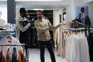 Shopping mall worker pointing at apparel rack while offering customer help in selecting casual outfit. Clothing store consultant assisting african american man in choosing garment photo