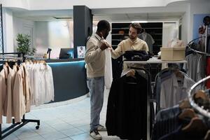 Clothing store customer holding shirt to body and asking fashion consultant for advice. African american man trying on formal apparel while shopping and getting help from boutique worker photo