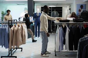 Stylish man searching for clothes and taking apparel from shelf in shopping mall department. African american shopper exploring apparel racks while choosing outfit in boutique photo