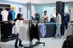 African american woman browsing rack with white shirts and selecting size in clothing store. Customer holding formal wear apparel and examining fit while shopping in fashion boutique photo