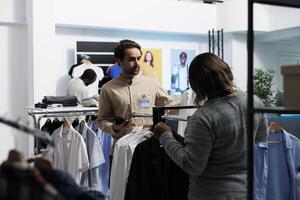 Clothing store worker showcasing shirt to customer and checking size option using digital tablet. Shopping center employee examining stock availability and helping client to choose apparel photo