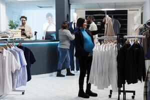 sonriente embarazada mujer participación chaqueta en percha mientras compras para maternidad ropa en Departamento centro comercial. expectante asiático mamá elegir hojeada estante y elegir atuendo en boutique foto