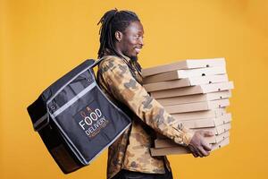 African american deliveryman carrying stack of pizza boxes, deliverying fast food orders to customers during lunch time. Restaurant employee standing in studio with yellow background. Food service photo