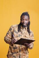 Confident person writing and taking notes on clipboard, checking papres with business job schedule in studio over yellow background. African american man looking at checklist, signing agreement files photo