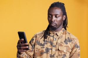 joven adulto mecanografía mensaje utilizando móvil teléfono, posando terminado amarillo antecedentes en estudio. africano americano hombre participación teléfono inteligente hojeada en Internet, leyendo nuevo en línea artículo foto