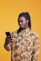 African american man reading good news on smartphone, scrolling through social media in studio over yellow background. Cheerful person searching information on phone, browsing on internet photo