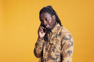 African american man talking with remote friend using mobile phone, chatting during studio shot. Excited young adult discussing about lifestyle, standing over yellow background photo