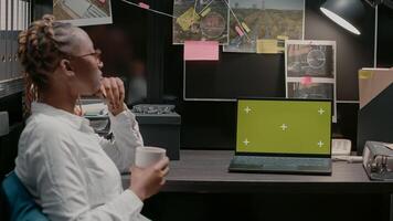 African american agent using greenscreen on laptop, analyzing isolated display and conducting criminal investigation. Female detective working with blank chroma key mockup screen. photo