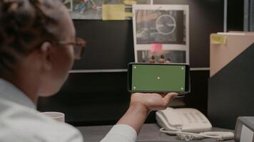 Police officer using greenscreen in evidence room, showing mockup display and conducting criminal investigation. Inspector checking isolated chroma key template with smartphone. photo