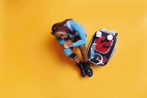 African american girl using mobile phone sitting on the floor in studio, having big trolley bag filled with travelling essentials next to her. Young adult with smartphone preparing to leave. photo